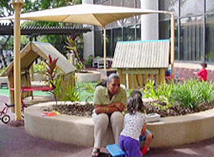 An adult sitting on a garden wall seat while a child plays on the wall next to her surrounded by playhouse structures and sun shades overhead on a child development center playground in Hawaii